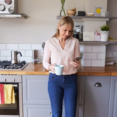 Woman Standing In Kitchen Sending Text Message On Phone